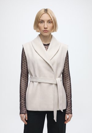 Donna con capelli biondi corti indossa un gilet beige senza maniche con cintura sopra una maglia nera trasparente a maniche lunghe con pois.