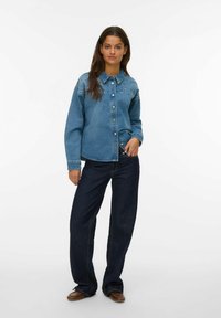 Camicia di denim azzurra con design a bottoni, caratterizzata da borchie decorative sulle spalle, abbinata a jeans larghi blu scuro.