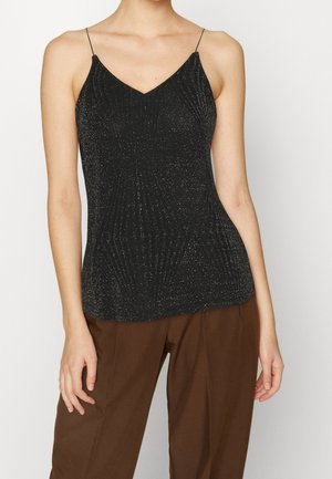 Femme portant un haut noir scintillant sans manches à col en V avec de fines bretelles et un pantalon marron, se tenant devant un fond uni.