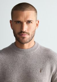Jasnoszary sweter z prążkowanym dekoltem i subtelną teksturą, z małym, wyszywanym logo na piersi. Portret zbliżeniowy.