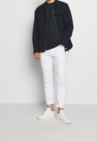 Námořnická zipová bunda přes černé tričko, doplněná o bílé slim-fit kalhoty a bílé tenisky. Hladké textury s minimalistickým designem.
