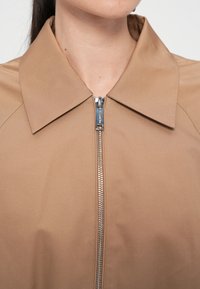 Ruda zip-up striukė su išsikišusiu apykakle, turi sidabrinę užtrauktuką ir lygų audinio paviršių. Nėra matomų raštų ar akcentų.