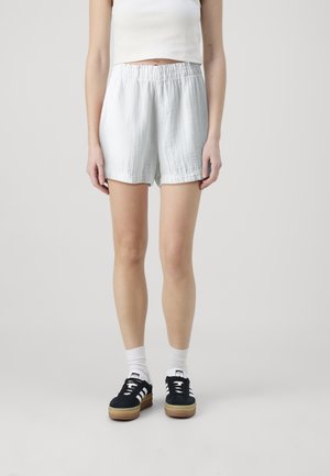 Personne portant un haut blanc sans manches et un short gris clair à taille élastique, avec des chaussettes blanches et des baskets noires à semelles marron.