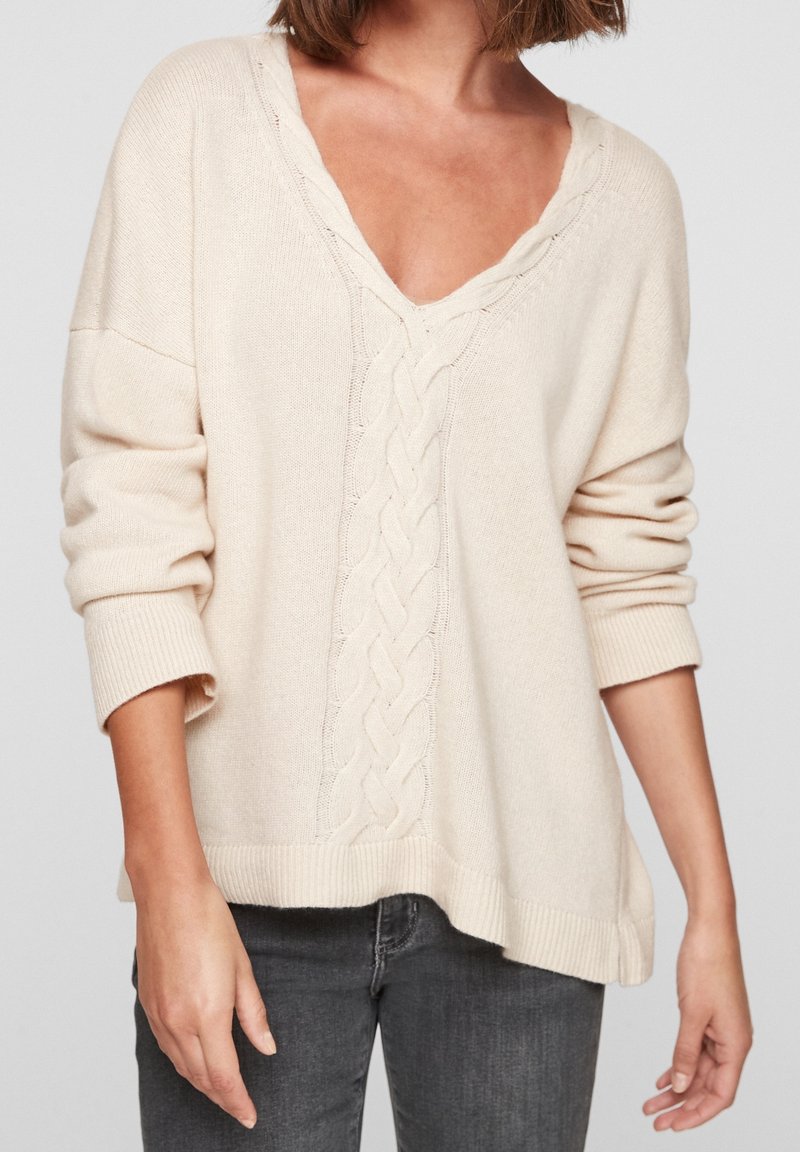 Femme portant un pull en tricot torsadé crème à col en V avec les manches retroussées et un jean gris foncé, sur un fond uni.