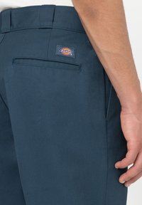 Detailní záběr ruky osoby odpočívající podél těla, oblečené v modrých kalhotách Dickies s kapsou na zadní straně a viditelnou logem.
