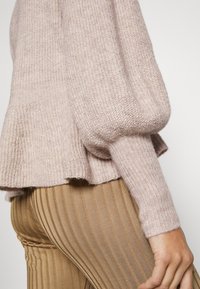 Primer plano de una persona que lleva un suéter de punto beige texturizado con mangas abullonadas y puños de canalé, combinado con pantalones color beige con rayas verticales.