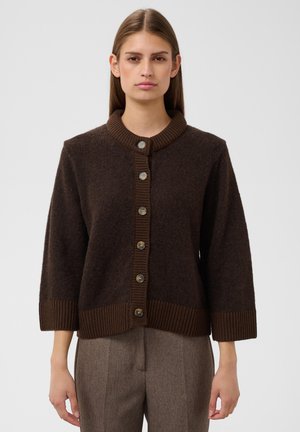 Jeune femme aux cheveux bruns lisses portant un cardigan marron foncé boutonné et un pantalon marron clair taille haute sur un fond uni.