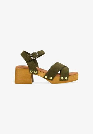Sandalia de ante verde oliva con tacón de bloque de madera, correa con hebilla en el tobillo, dos tiras delanteras y detalles de tachuelas doradas a lo largo de la suela.