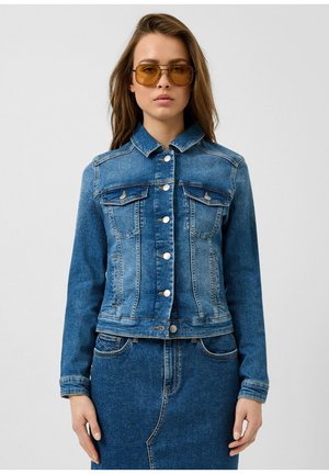 Vrouw met amberkleurige zonnebril, een blauwe spijkerjack met knoopsluiting en een bijpassende spijkerrok tegen een effen grijze achtergrond.