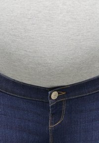 Lähikuva henkilöstä, joka pukeutunut harmaaseen paitaan ja jonka harmaat farkut ovat napittamatta.