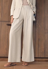 Bež hlače širokih nogavica s okomitim prugama, srednje teške tkanine i bočnim džepovima. Uparen s ravnim, svijetlim sandalama.