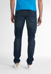 Tmavě modré denimové džíny s rovný střihem, obsahující zadní kapsy a jemné vyblednutí. Nosené s bílými teniskami s hnědým akcentem.