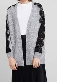 Grijze gebreide cardigan met lange zwarte kanten mouwen, open voorkant en geribbelde manchetten, gecombineerd met een wit topje en een zwarte rok.