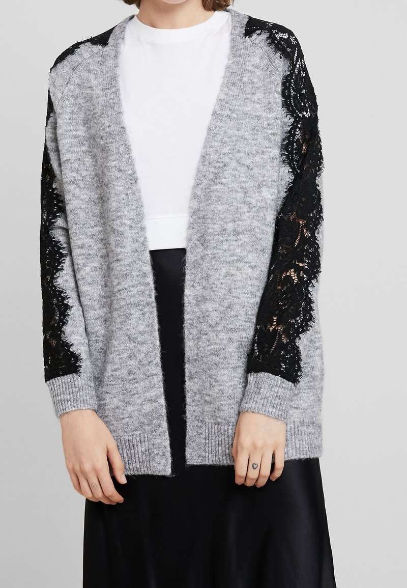 Grijze gebreide cardigan met lange zwarte kanten mouwen, open voorkant en geribbelde manchetten, gecombineerd met een wit topje en een zwarte rok.