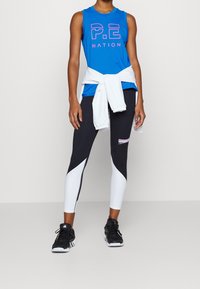 Top sin mangas azul con el logo "P.E NATION", sudadera blanca atada a la cintura, legging negros con acentos blancos y grises, zapatillas negras.