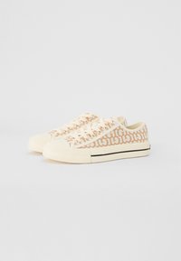Beige canvas sneakers med ett mönstrat övre, med vita snören, gummitåhattar och ett svart band längs sulan.