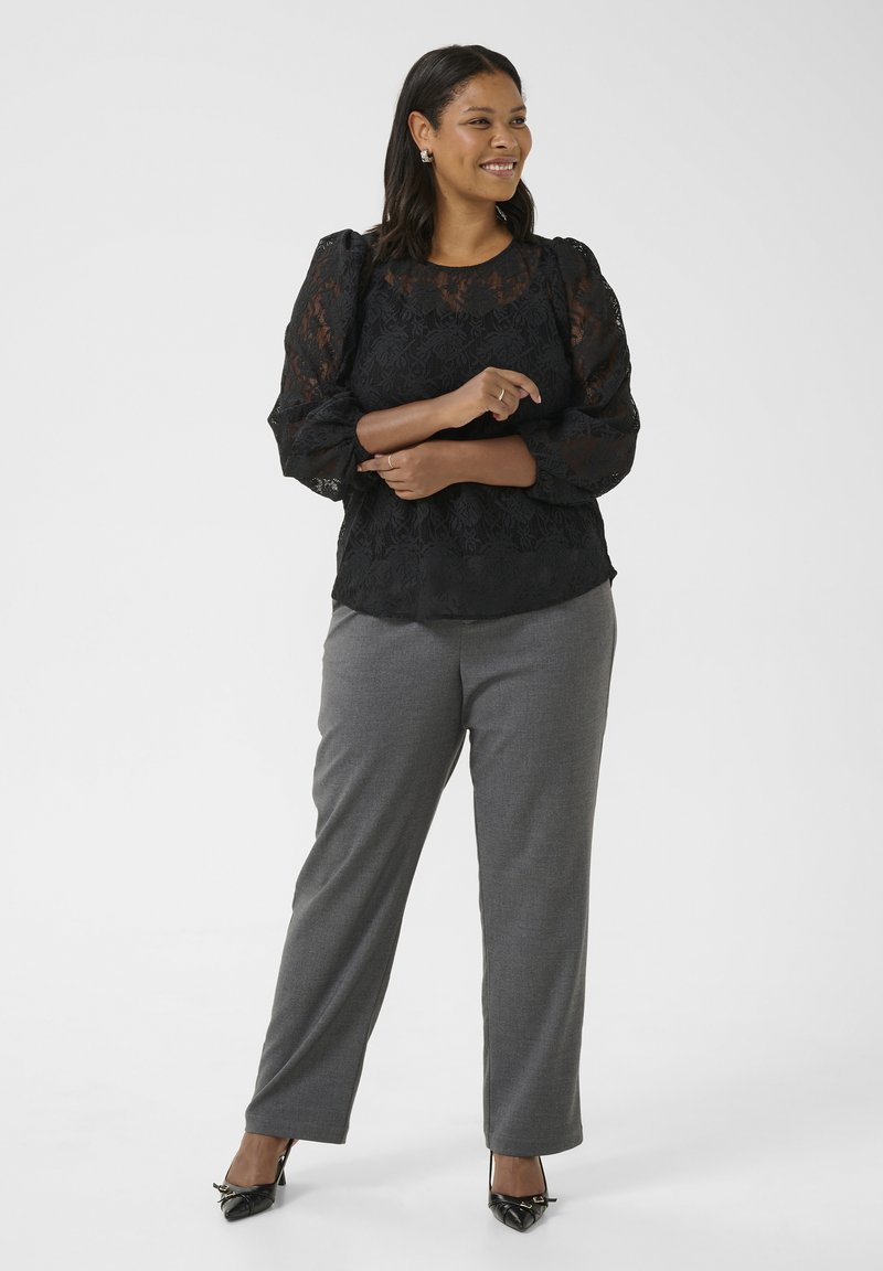 Blouse en dentelle noire avec des manches bouffantes, associée à un pantalon gris ajusté. Le mannequin porte des chaussures à talons noirs pointus et des bijoux en argent.