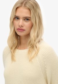 Donna bionda con onde morbide, che indossa un maglione lavorato a maglia color crema, guarda leggermente oltre la fotocamera su uno sfondo bianco.