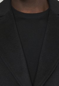 Primo piano di una persona che indossa un maglione a girocollo nero sotto un blazer di lana scura.