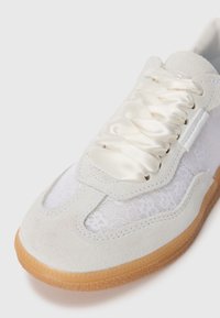 White sneaker with suede and lace textures, cream satin laces, and a gum rubber sole. Features a smooth, rounded toe and stitched accents.