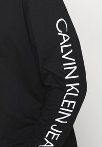 Černá košile s dlouhými rukávy, na které je na pravém rukávu nápis "CALVIN KLEIN JEANS" v tučném bílém písmu. Vyrobena z hladké tkaniny a má volný střih.