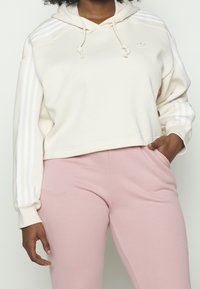 Uppskuren beige hoodie med vita snören och tre vita ränder på ärmarna, parat med rosa byxor. Mjuk tyg, avslappnad passform.