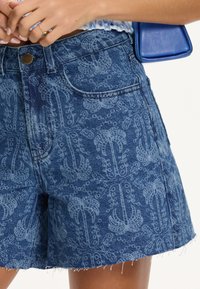 Shorts en denim bleu moyen avec un léger motif floral. Dotés d'une taille haute, d'un ourlet effiloché et de poches avant. Agrémentés d'un bouton en laiton.