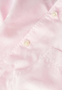 Camisa rosa claro con una textura suave, que cuenta con un collar doblado, cierre de botones y un bolsillo en el pecho. Detalle de botón notable visible.