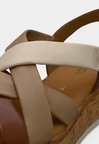 Gros plan d'une sandale compensée avec des brides en cuir beige, taupe et marron superposées et une semelle texturée liège.