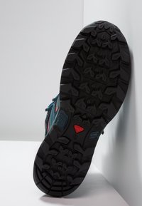 Trailová bota s černou gumovou podrážkou s hlubokými vzory pro lepší přilnavost, vršek z tyrkysové tkaniny s červenými detaily a šněrováním.