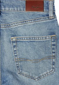 Gaiši zili džinsi ar aizmugures kabatu, kurai ir dzelteni diegi un brūns ādas logotipa uzšuvums ar uzrakstu "Pepe Jeans London".