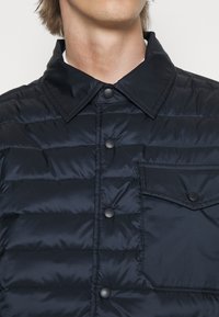 Giacca a vento navy con pannelli cuciti, colletto a punta, tasca frontale e bottoni a pressione neri per la chiusura. Texture liscia e riflettente.