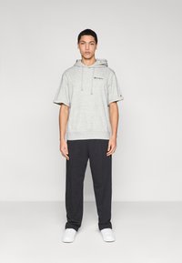 Γκρι κοντομάνικο φούτερ με κουκούλα και ρυθμιζόμενο κορδόνι, με λογότυπο. Συνδυασμένο με σκούρο φόρμα παντελόνι και λευκά αθλητικά παπούτσια. Απλό φόντο.