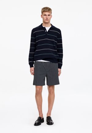 Man draagt zwarte loafers, grijze kortebroek met krijtstrepen, wit onderhemd en zwarte trui met witte en groene horizontale strepen, staande op een witte achtergrond.