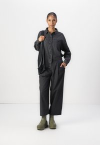 Camisa de botão em denim escuro combinada com calças largas. Modelo segura uma bolsa preta, vestindo botas de plataforma verdes.