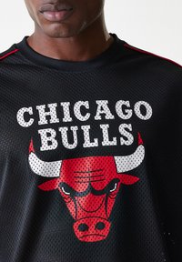 Melns sieta sporta krekls ar uzrakstu "CHICAGO BULLS" baltā krāsā, ar sarkanu bulli zem tā. Akcenti ietver sarkanas svītras uz pleciem.