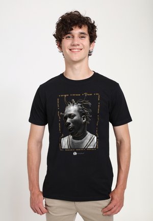 Ung mand smiler, iført en sort T-shirt med en sort-hvid grafik af en person med fletninger og tekst med gyldne konturer.