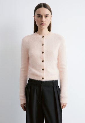 Kobieta o prostych brązowych włosach, nosząca złote kolczyki-koła, jasnoróżowy, miękki kardigan na guziki oraz czarne spodnie z wysokim stanem, stojąca na tle białej ściany.