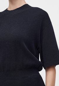 Femme portant un haut en maille noir à manches courtes avec un col rond montant et des poignets et ourlet côtelés.
