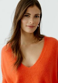 V-Ausschnitt, übergroßer Pullover aus leuchtend orangefarbenem Strick, mit weichem Material und lockerer Passform, sowie einem dezenten Rippensaum am Ausschnitt.