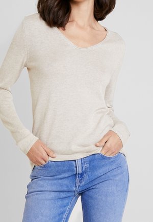 Femme portant un pull beige clair à manches longues avec un col en V, légèrement rentré dans un jean bleu moyen, les mains dans les poches.