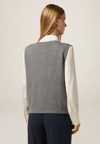 Suéter sin mangas gris, textura de punto, escote redondeado, usado sobre una camisa blanca de satén, con pantalones azul marino de corte suelto visibles.