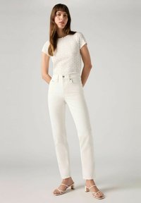 Witte top met korte mouwen en blauwe bloemenpatroon, gecombineerd met hoge witte jeans en witte sandalen met een blokhak.