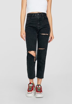 Femme portant un jean noir déchiré à taille haute, raccourci aux chevilles, associé à des baskets blanches avec des touches rouge et vert et des lacets multicolores.