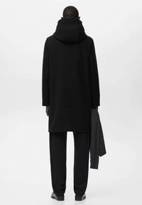 Abrigo de lana negro con capucha alta, forma recta y mangas largas. Una bufanda gris cuelga sobre el brazo izquierdo. La modelo está de espaldas al espectador.