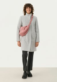Helle graue Wolljacke mit hohem Kragen, kombiniert mit schwarz gestreiften Hosen und kräftigen schwarzen Stiefeln. Eine pinke Gürteltasche ist schräg über die Schulter getragen.