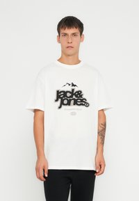 Camiseta de algodón blanca con un gran logo negro y gris que presenta gráficos de montañas. Mangas cortas y cuello redondo. Diseño simple y casual.