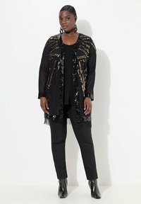 Kimono negro con lentejuelas y detalles de flecos, que presenta patrones lineales dorados. Lleva puesto sobre una blusa negra y se combina con pantalones negros y botas de tobillo.