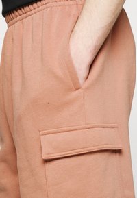 Primer plano de la mano de una persona en el bolsillo lateral de unos pantalones deportivos color durazno con un bolsillo adicional con solapa justo debajo.