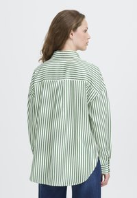 Donna di profilo che indossa una camicia a maniche lunghe a righe verticali verdi e bianche e jeans blu, su uno sfondo neutro.